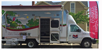 The Bookmobile at a stop in front of a house. The side door of the bookmobile is open and the canopy is extended. 